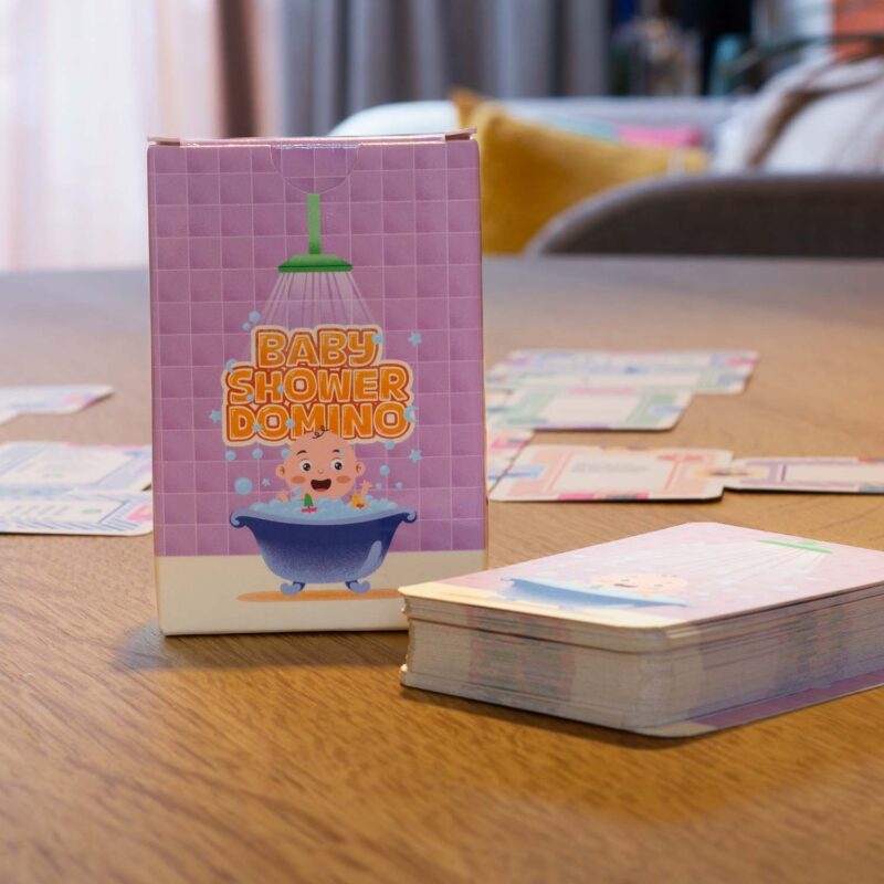 Het Babyshowerdomino spel klaargezet op een tafel in een gezellige woonkamer, met het bewaardoosje rechtop en een stapel speelkaarten ernaast.